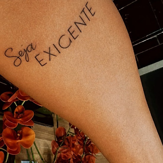 A frase 'Seja Exigente' tatuada na perna, com orquídeas na composição