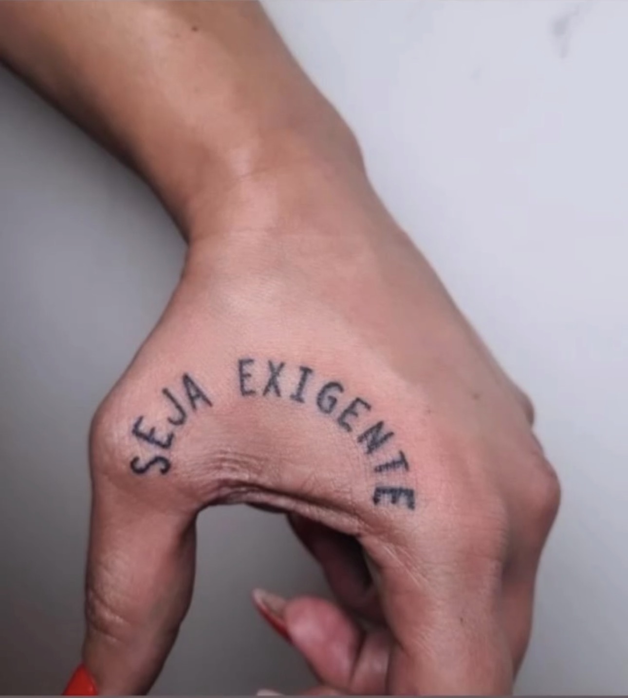 'Seja Exigente' tatuado em arco no dorso da mão