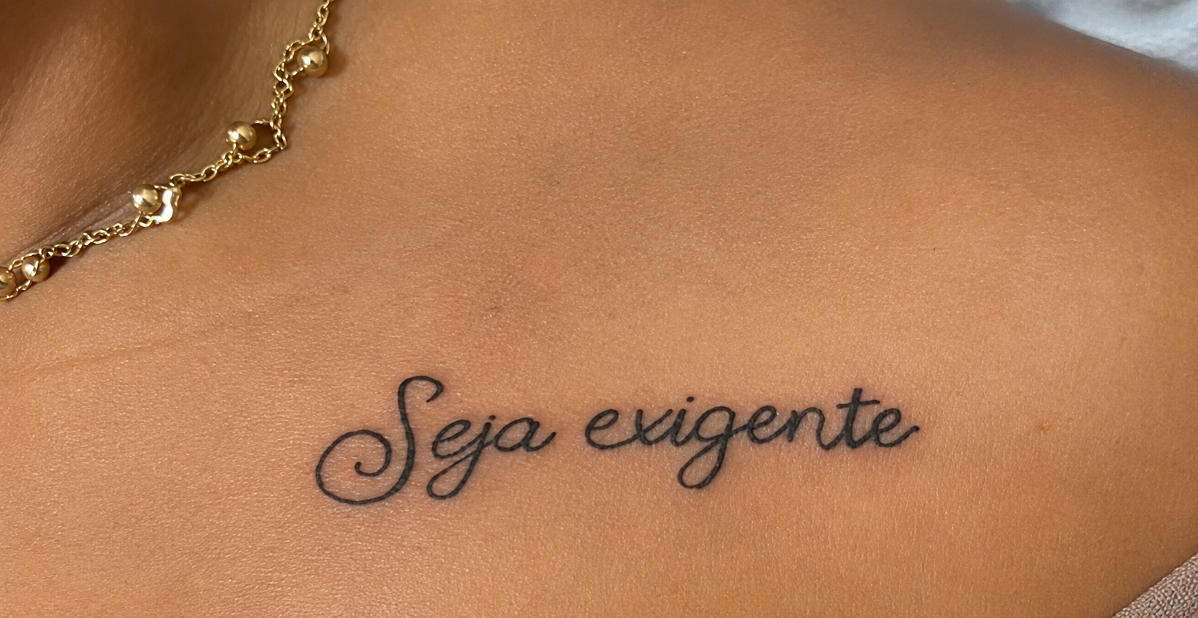 'Seja exigente' em caligrafia fina no colo, com o colar dourado acima
